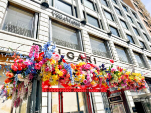 Colorful floral installation hanging above the entrance to the Vera Wang boutique, with bright flowers in pink, blue, yellow, and white against the building’s exterior.