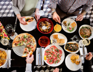 Overhead view of a table filled with Italian dishes, including pizza, pasta, focaccia, salads, cocktails, and wine, with several people sharing the meal.