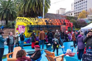 A colorful food truck serving a crowd in Union Square, surrounded by people enjoying outdoor seating under tall palm trees.