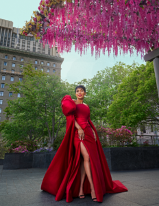 A woman poses in a red floor length gown, with pink flowers overhead