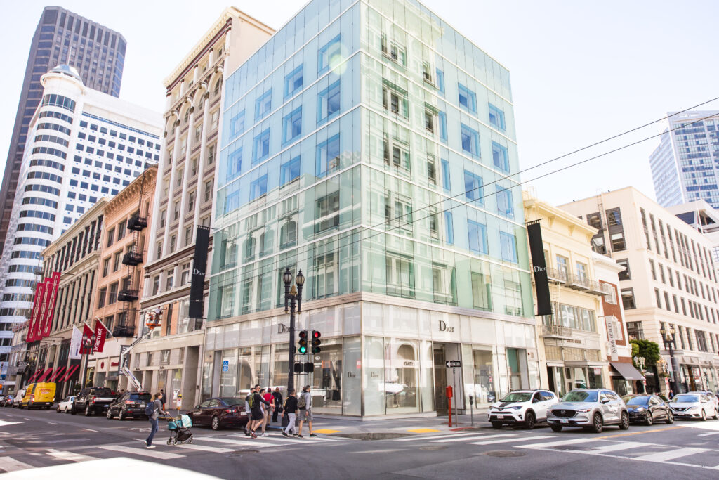 Modern glass-clad Dior retail store on a street corner in a downtown shopping district, surrounded by multi-story buildings, pedestrians crossing the intersection, and parked cars in Union Square, San Francisco